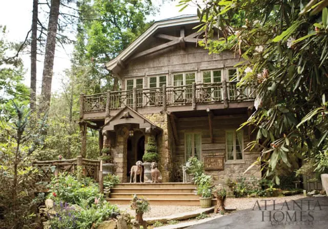 Rustic log and stone mountain home with a wooden porch and balcony, surrounded by lush trees and foliage. Two dogs sit on the front steps.