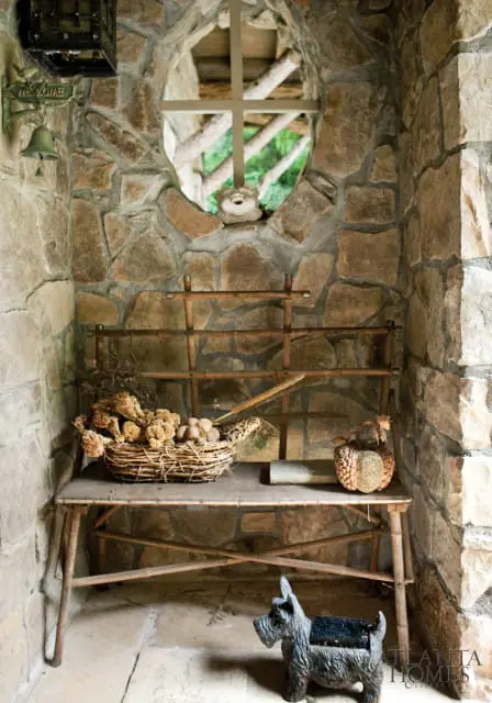 Stone alcove with a rustic wooden bench holding a basket of dried flowers and a decorative gourd, beneath an oval window. A Scottie dog statue sits on the stone floor.