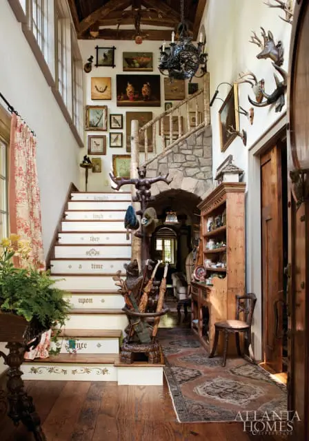 Rustic entryway with a stone archway leading to a hallway, a wooden staircase with painted phrases on each riser, and walls adorned with numerous framed artworks and taxidermied deer heads.