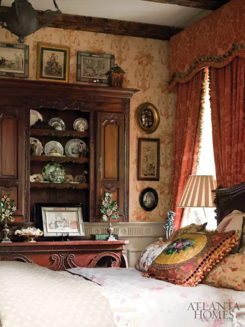 Bedroom with floral wallpaper, antique wooden hutch filled with plates, and ornate red dresser. A large window is draped with coral curtains.