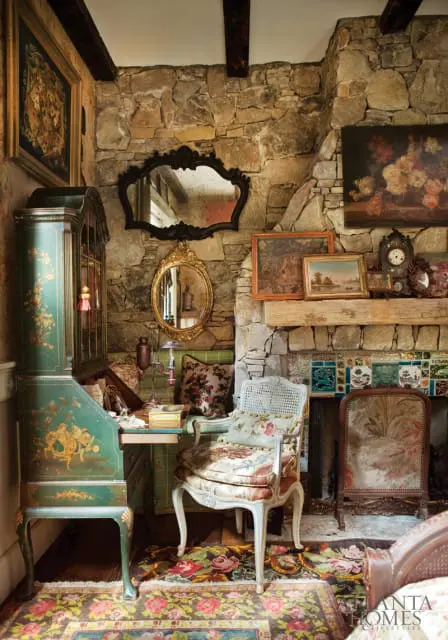 Eclectic living room with stone walls, a green painted secretary desk, floral upholstered chairs, and a fireplace with decorative tiles.