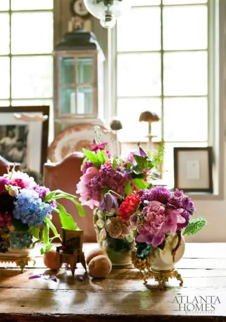 A rustic wooden table displays three vases filled with colorful peonies, hydrangeas, and other blooms, alongside peaches and decorative objects.