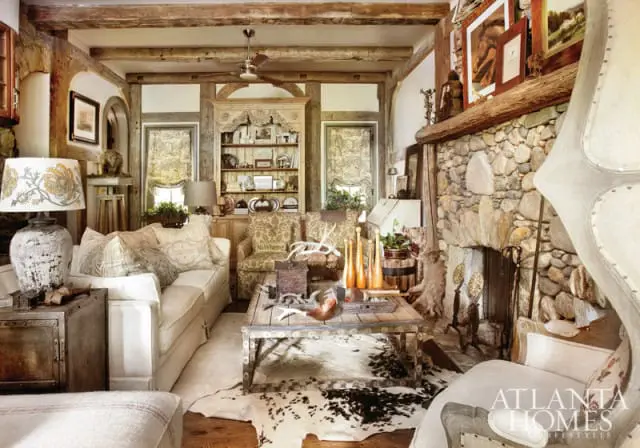 Rustic living room with exposed wooden beams, a large stone fireplace, a white sofa, patterned armchairs, and a distressed wood coffee table on a cowhide rug.