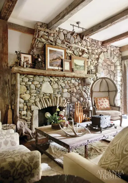 Living room with a large stone fireplace, exposed wooden beams, a rustic wooden mantel with framed art, and a patterned armchair.