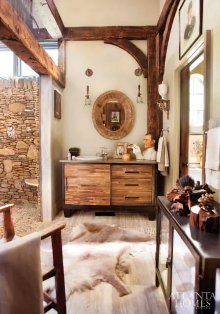 Rustic bathroom with stone shower, wooden vanity with slatted drawers, oval wood mirror, and cowhide rugs on wood-look tile floor.