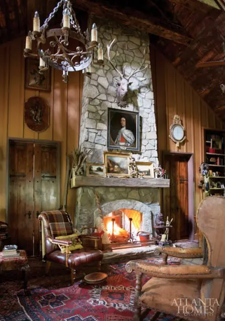 Rustic living room with a large stone fireplace, mounted deer head, oil paintings, wood-paneled walls, and antique furniture.