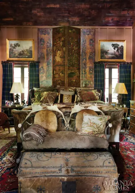 Bedroom with a rustic, maximalist design featuring a fur-covered bed adorned with antlers and animal print pillows, a large decorative screen, plaid curtains, and antique trunks.