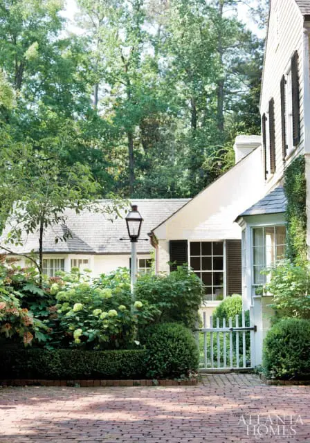 White brick house with black shutters and a white picket gate, surrounded by lush green landscaping and a red brick driveway.