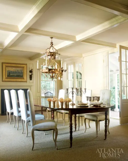 Dining room with coffered ceiling, ornate chandelier, fireplace, and a long table set for a meal with white upholstered chairs.