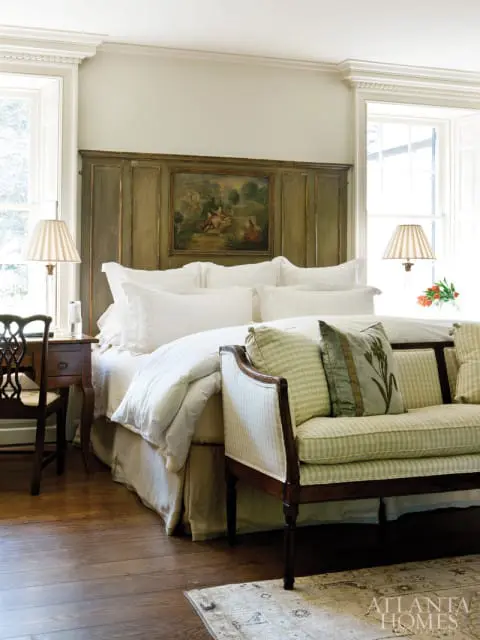 Bedroom with a large bed dressed in white linens, a distressed green paneled headboard with a small landscape painting, and a striped settee.