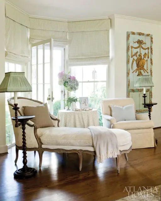 Living room with a chaise lounge, armchair, and round table covered in a gingham tablecloth. Light green gingham Roman shades cover French doors and windows.