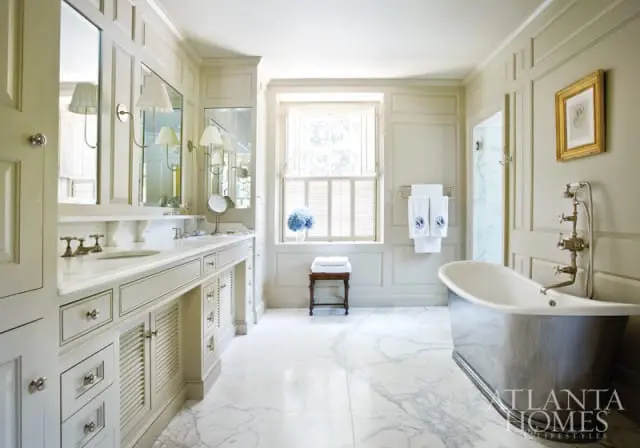 Cream bathroom with marble floors, double vanity with white countertop, freestanding tub with chrome fixtures, and a window with white shutters.
