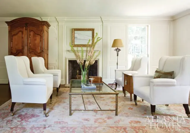 Living room with white upholstered armchairs, a large wooden armoire, a fireplace with artwork above, and a glass-top coffee table with a floral arrangement.