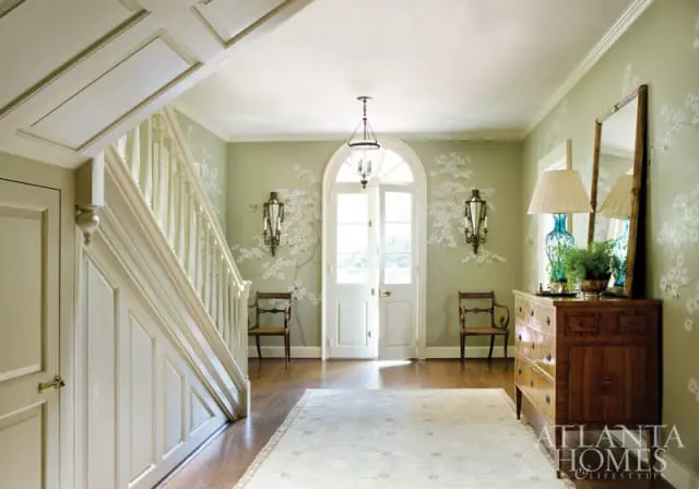 Foyer with light green walls featuring a cherry blossom mural, a grand staircase with white paneling, a large arched doorway, and a wooden dresser topped with a lamp and plants.