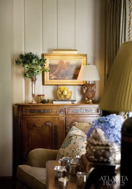 Antique wooden sideboard with ornate carvings, topped with a painting, vase of greenery, bowl of pears, and a lamp. Upholstered furniture and floral pillows are in the foreground.