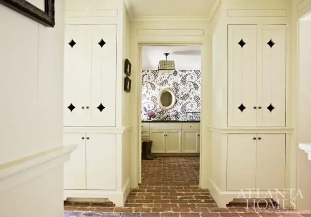 Hallway with cream-colored built-in cabinetry featuring diamond-shaped cutouts and brass hardware, leading to a room with patterned wallpaper, a vanity, and a plaid pendant light.