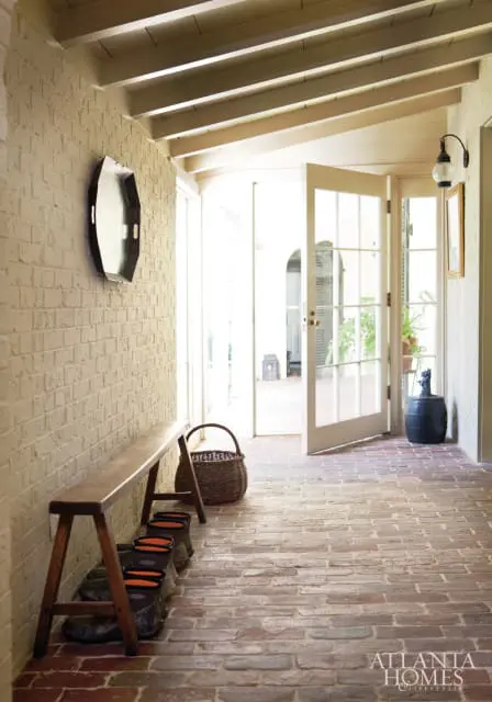 Hallway with white brick walls, a wooden bench with boots underneath, and a brick floor. Double doors open to a sunlit courtyard.