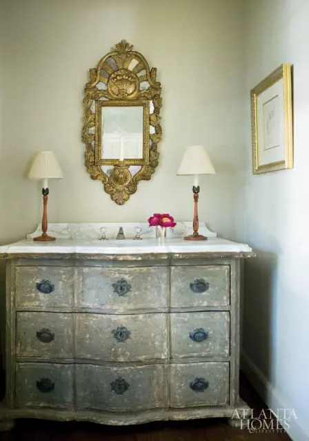 A distressed blue dresser with a marble top topped with two lamps, a mirror, and a vase of pink flowers.