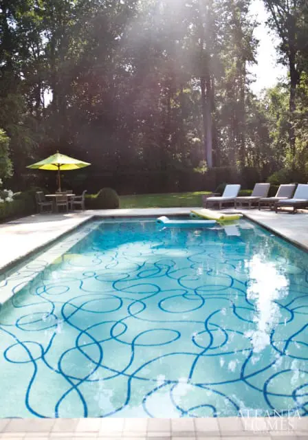 Rectangular swimming pool with blue swirling tile pattern, surrounded by a stone patio, lounge chairs, and lush trees.
