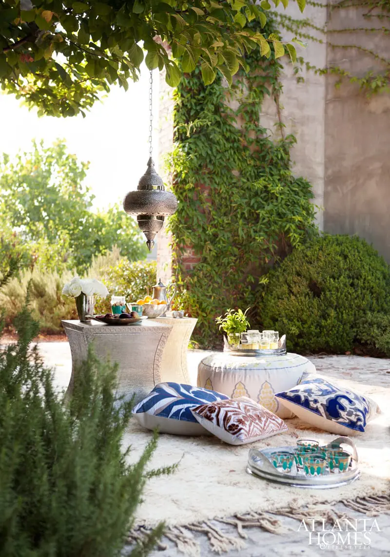 Outdoor seating area with a silver Moroccan lantern hanging above a textured table with white roses and fruit, surrounded by patterned floor cushions and a plush rug.