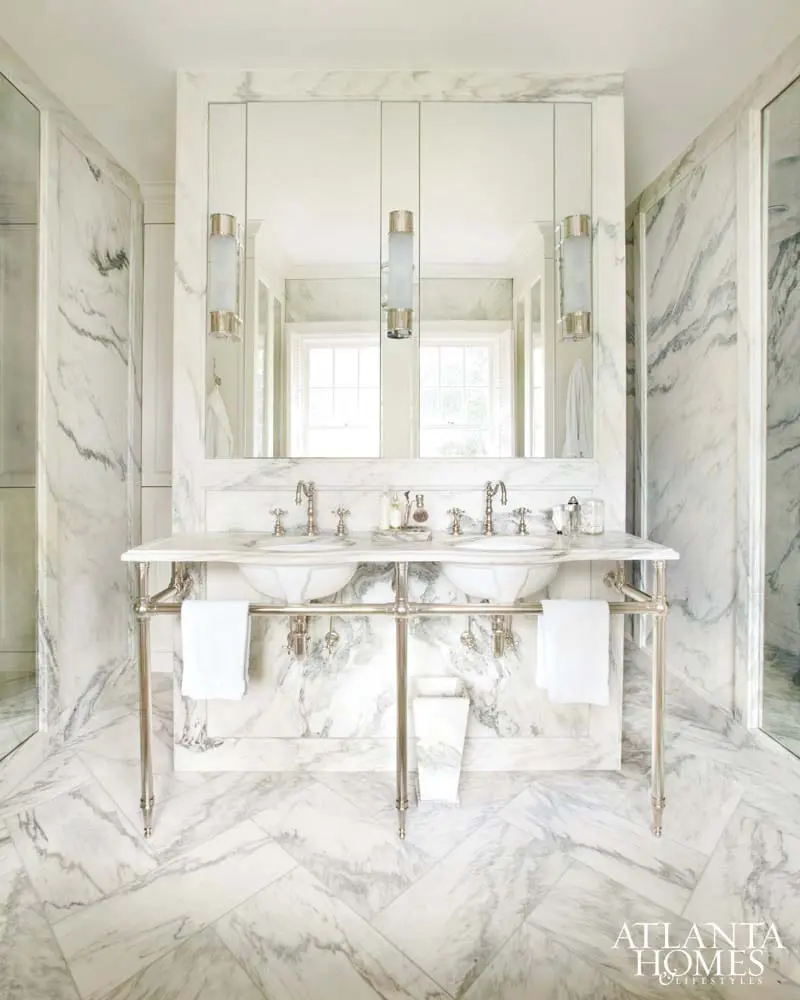 Marble bathroom with a double vanity featuring undermount sinks, polished nickel fixtures, and a mirrored medicine cabinet. The floor is laid in a herringbone pattern of marble tiles.