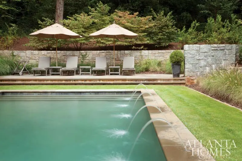 Swimming pool with fountains, lounge chairs, and umbrellas on a wooden deck against a stone wall and lush greenery.