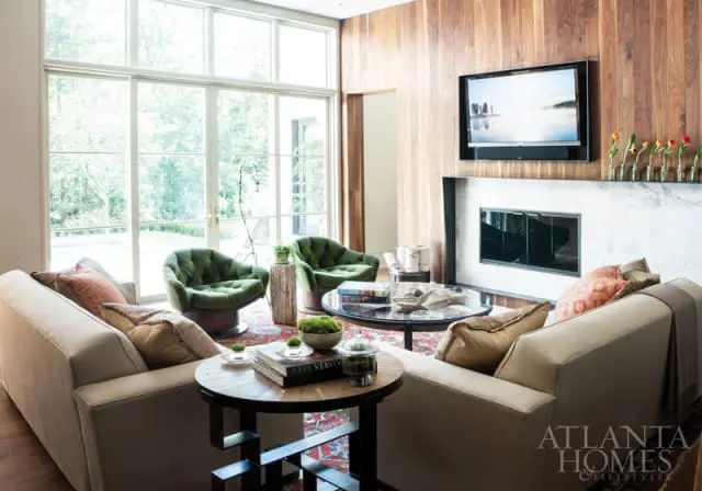 Living room with floor-to-ceiling windows, wood-paneled walls, a marble fireplace, two beige sofas, two green velvet armchairs, and a glass-top coffee table.