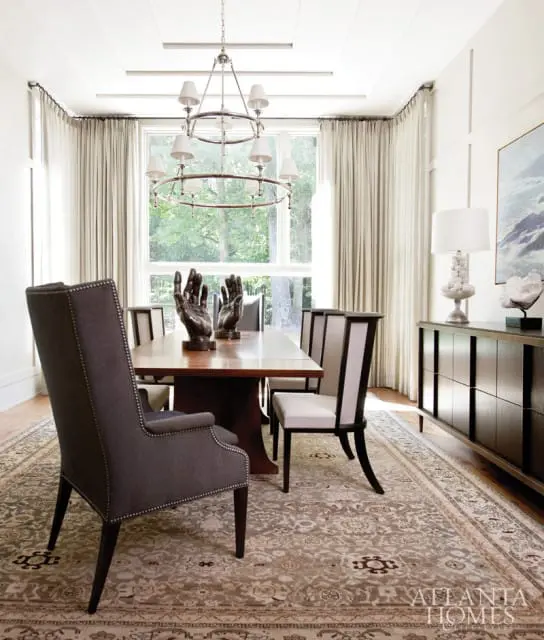 Dark gray upholstered dining chairs with nailhead trim surround a wooden dining table topped with hand sculptures. A large window with sheer curtains and a modern chandelier are visible.