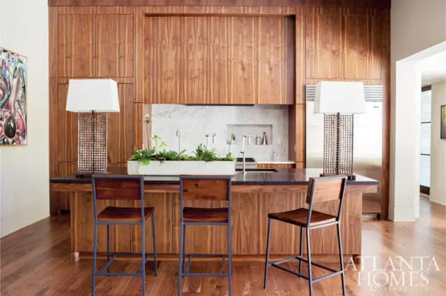 Modern kitchen island with dark granite countertop, warm wood cabinetry, and three wooden bar stools. Marble backsplash with faucets and a concrete planter filled with greenery.