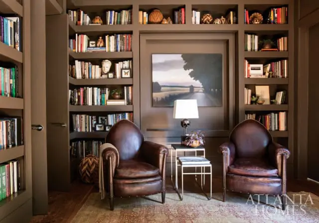 Brown leather club chairs flank a side table with a lamp and plant, in front of floor-to-ceiling bookshelves filled with books and decorative objects.
