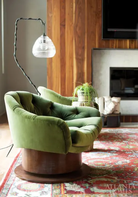 Green velvet tufted swivel chairs in a living room with a fireplace, wood-paneled wall, and oriental rug.