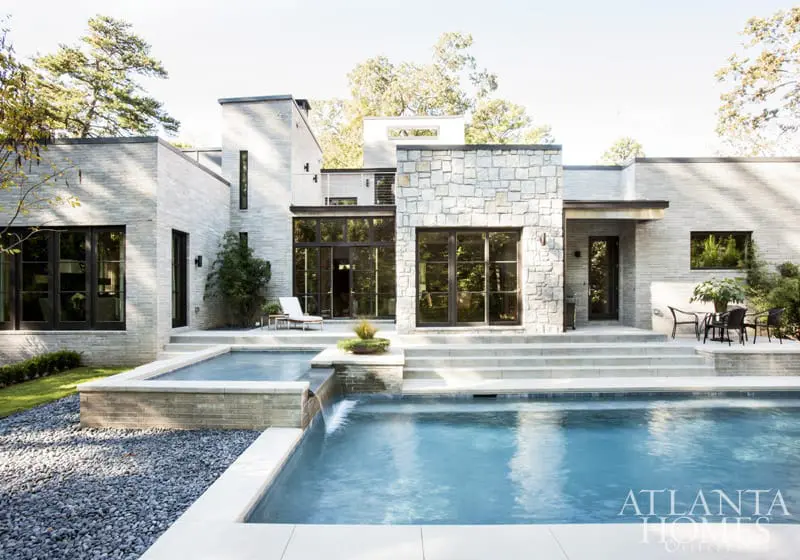 Modern home with light grey brick and stone facade, featuring large dark-framed windows, a multi-level swimming pool, and a stone patio with outdoor seating.