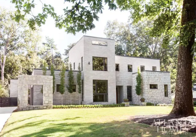 Modern home with light stone facade, large dark-framed windows, and a stone entryway structure. Lush green lawn and trees surround the property.