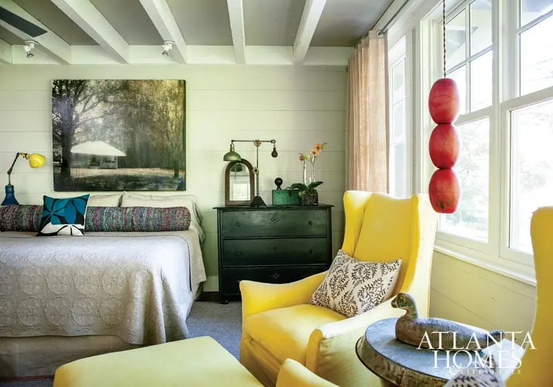 Bedroom with a light green shiplap wall, white beamed ceiling, and a large abstract painting above the bed. Features include a dark green dresser, yellow armchair, and a stack of red buoys hanging in the window.