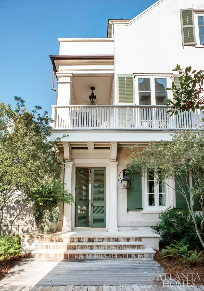 White stucco house with green shutters and double doors, white balcony railing, and lush landscaping.