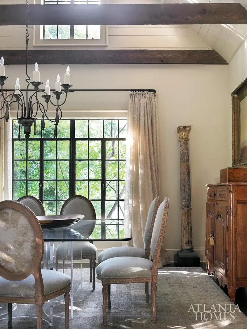 Dining room with a glass-top table and upholstered chairs, a dark metal chandelier, and a wooden cabinet. Large windows reveal a lush green outdoor view.