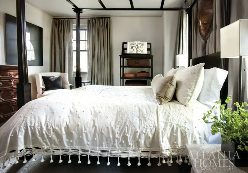 Bedroom with a four-poster bed dressed in white embroidered bedding with tassels, a cream armchair, and dark wood furniture.