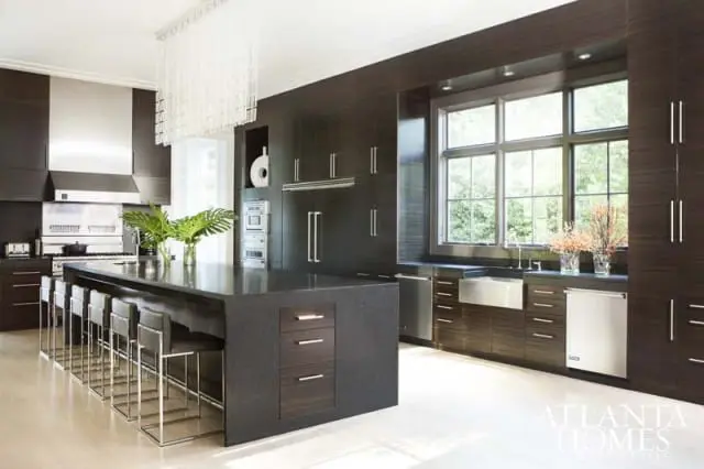 Dark wood kitchen with a large granite island, stainless steel appliances, and a large window overlooking greenery.