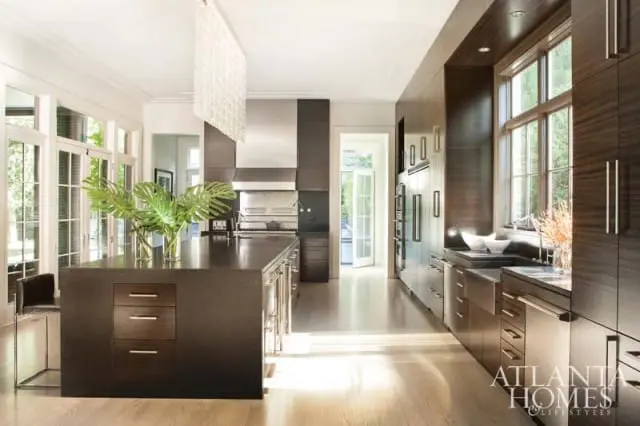 Dark wood kitchen with a large island, stainless steel appliances, and a long counter space with a sink. Large windows and glass doors let in natural light.