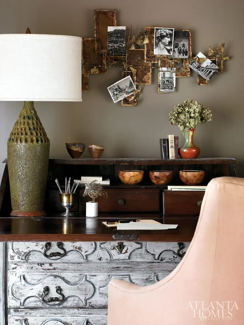 Antique wooden secretary desk with distressed white drawers, featuring a large green textured lamp, metal wall art with attached black and white photos, and a coral-colored vase with dried flowers.