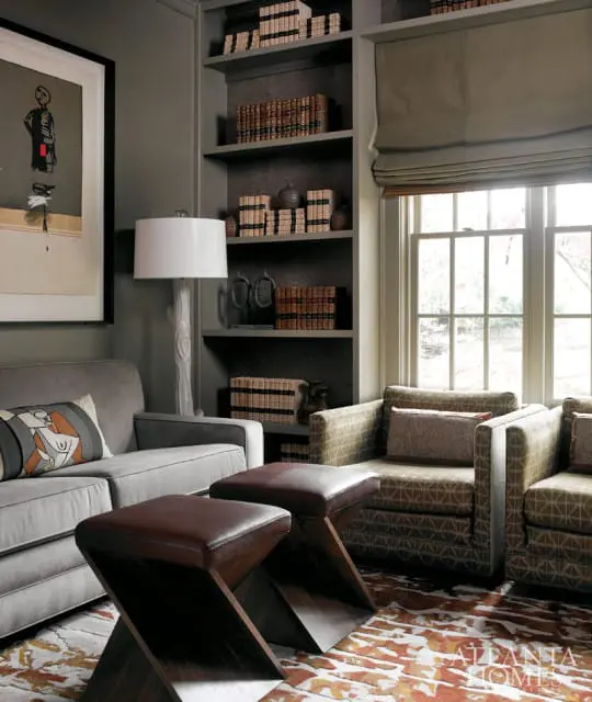 Gray living room with built-in bookshelves filled with books, a gray sofa, patterned armchairs, leather ottomans, and a large abstract artwork.