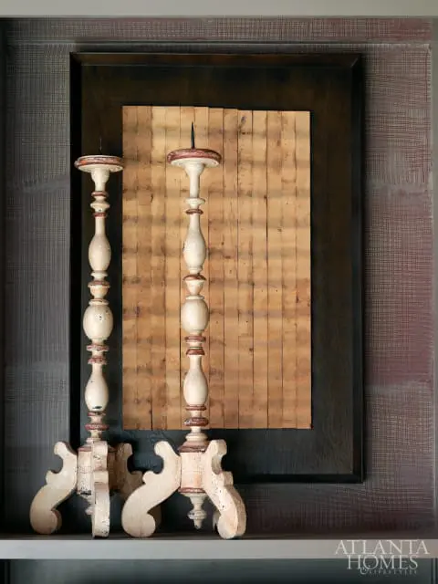 Two distressed antique wooden candlesticks with red and white accents stand in front of a framed piece of wood paneling and textured wallpaper.