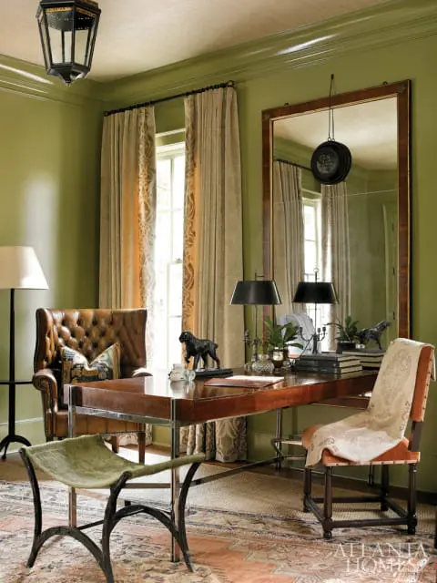 Office with olive green walls, a tufted leather wingback chair, a leather-topped desk with metal frame, and a decorative rug.
