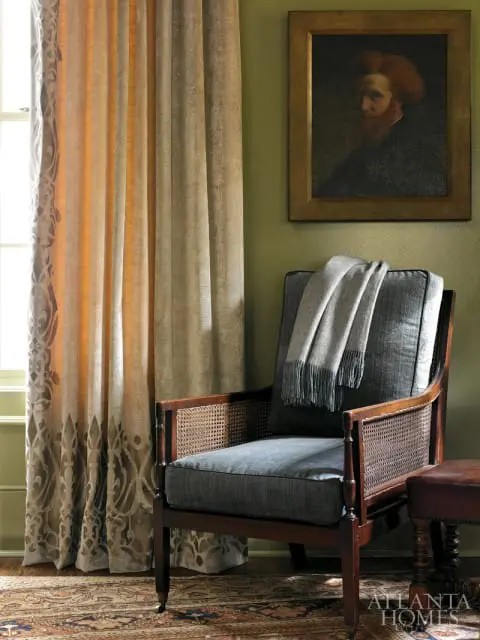 Dark wood armchair with cane sides and upholstered seat, draped with a gray throw, next to patterned curtains and a framed portrait on a green wall.