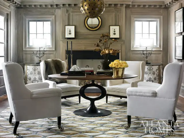 Living room with paneled wood walls, a round wooden table with four white armchairs, a fireplace, and a large geometric rug.