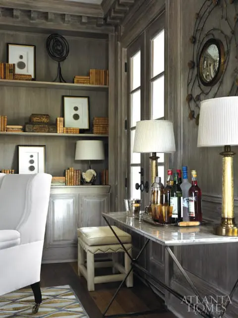 Gray wood-paneled study with built-in bookshelves filled with books and framed art, a marble-topped bar cart with bottles and glassware, and a white upholstered armchair.