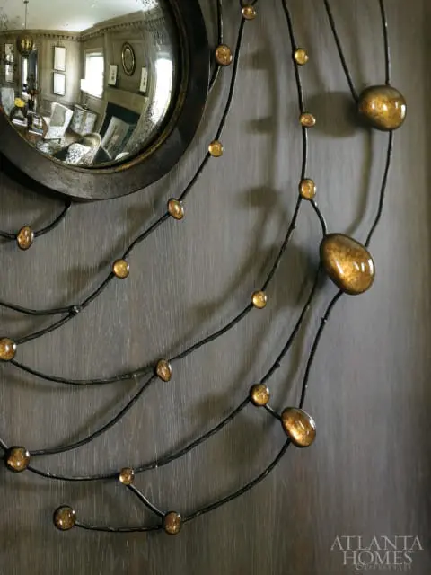 Close-up of a dark metal wall sculpture with amber glass orbs, reflecting a room with a fireplace and furniture.