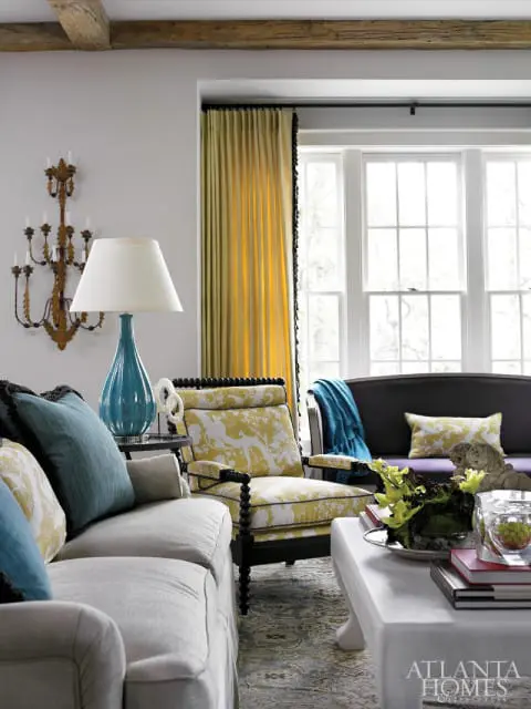 Living room with a light gray slipcovered sofa, a yellow floral armchair, and a dark sofa with a blue throw. Yellow curtains hang over a large window.