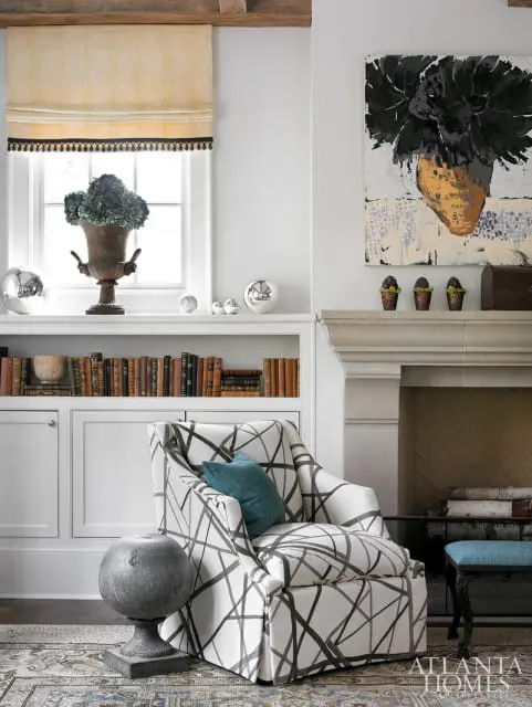 Living room with a white armchair featuring a black abstract pattern, a teal throw pillow, built-in white shelving with books, and a fireplace with stone surround.