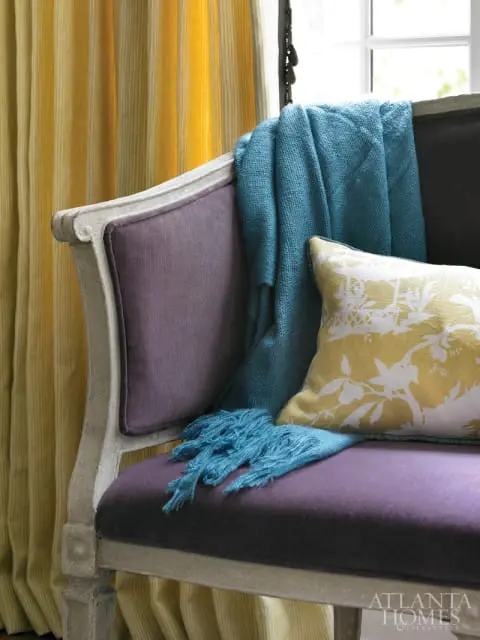 Purple upholstered bench with a teal throw blanket and a yellow floral pillow, next to yellow striped curtains.
